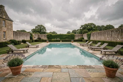Piscine extérieure d'un hôtel de charme en Normandie, entourée de verdure et de murs en pierre normande sous un ciel nuageux