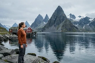 Femme en pull en laine contemplant les montagnes et les fjords des îles Lofoten en Norvège, avec des cabanes de pêcheurs rouges traditionnelles en arrière-plan